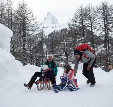 Mum sitting on a toboggan with one kid, while dad pushes the other child on their own toboggan | © Michael Portmann